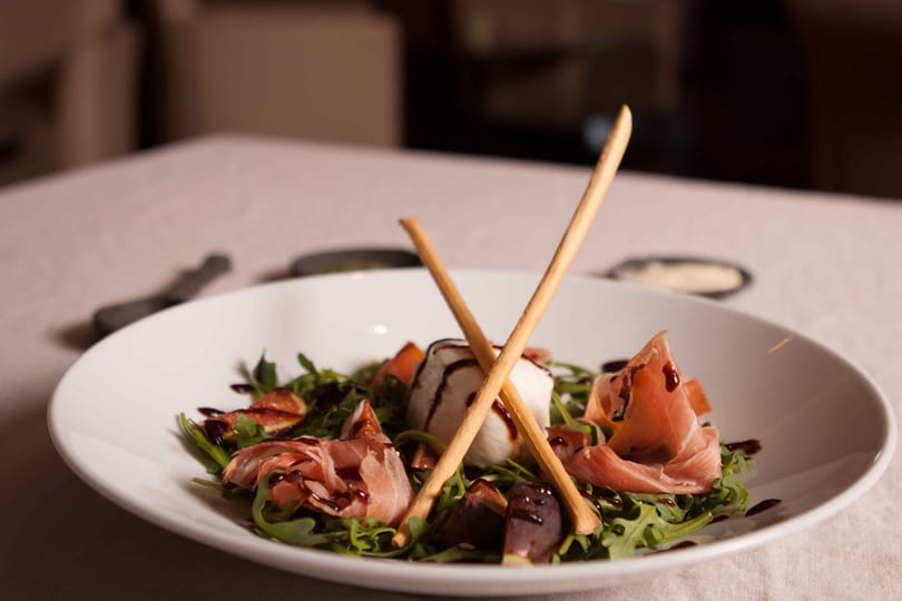 Arugula salad with prosciutto, figs, mozzarella, balsamic glaze, and breadsticks in a white bowl.