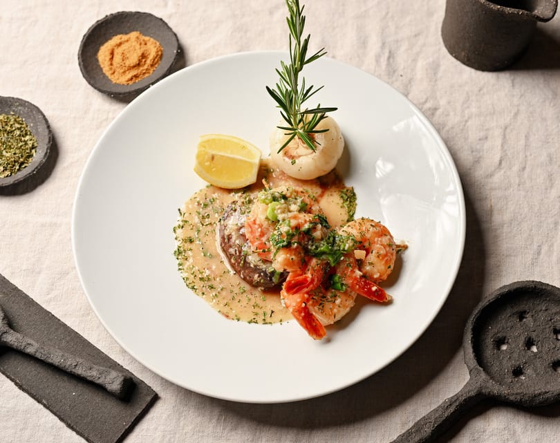 Gourmet dish with shrimp, steak, lemon, and a mushroom garnished with rosemary on a white plate.