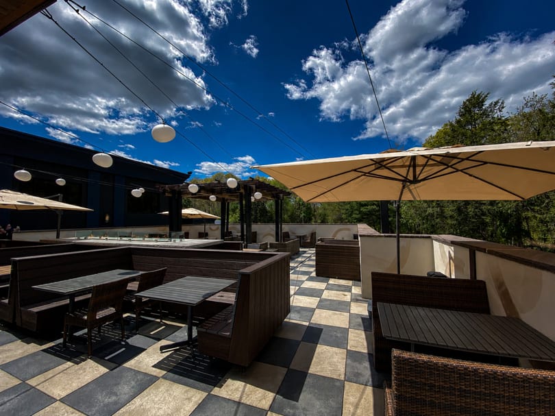 Outdoor patio with checkered floor, dark wooden booths, beige umbrellas, and string lights under a blue sky.