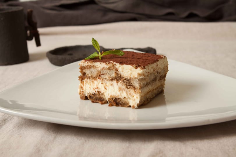 A slice of tiramisu with cocoa powder and a mint leaf on a white plate.