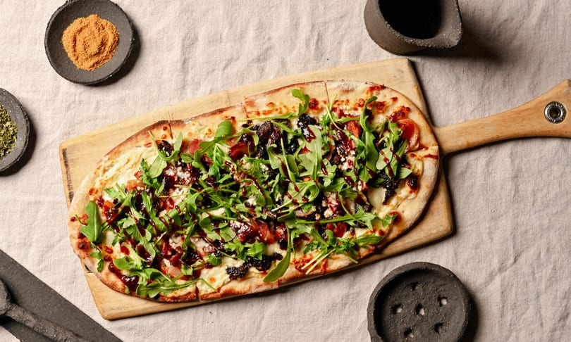 Flatbread pizza with arugula, cheese, bacon, figs, and balsamic glaze on a wooden board.