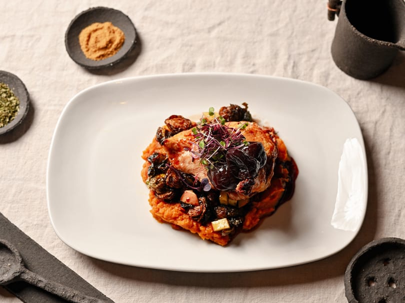A gourmet dish of chicken with dark fruit and microgreens on a bed of sweet potato mash.