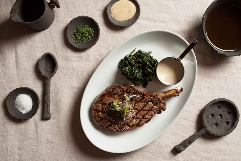 Grilled steak with greens, sauce, and seasonings on a white plate, surrounded by dark ceramic dishes.