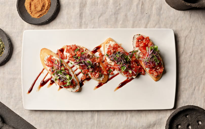 Five bruschetta pieces with diced tomatoes, microgreens, and balsamic glaze on a white platter.